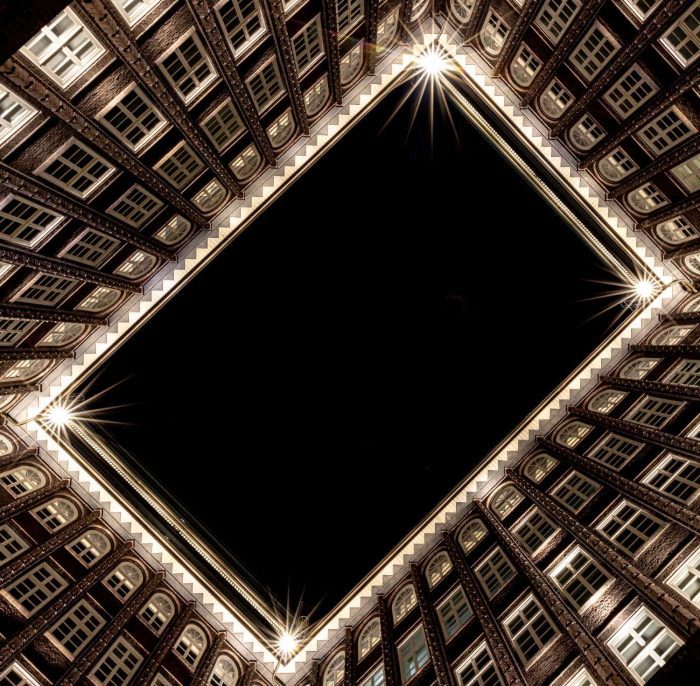 Blick nach oben aus der Mitte eines quadratischen Innenhofs, umgeben von mehrstöckigen Backsteingebäuden mit vielen Fenstern. Helle Lichter erhellen jede Ecke und rahmen den dunklen Nachthimmel darüber ein.