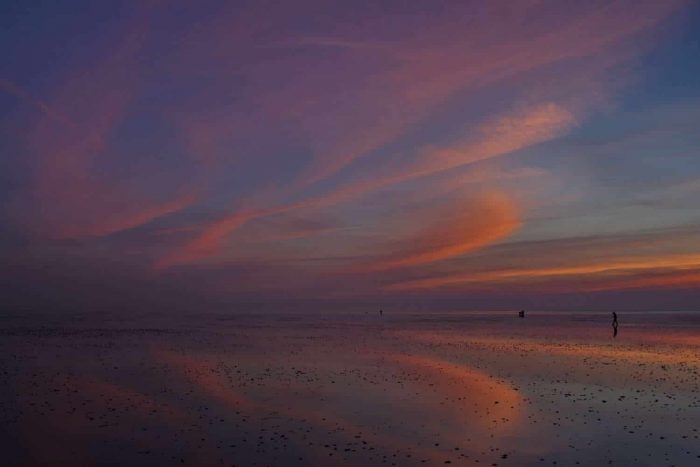 Eine weite Strandszene bei Sonnenuntergang mit leuchtend orangefarbenen, rosa und violetten Wolken, die sich im nassen Sand spiegeln, und ein paar Menschen, die in der Ferne am Ufer entlanggehen.