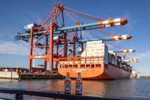 Zwei Bierflaschen auf einem Geländer, im Hintergrund Unscharf ein Containerschiff mit Hafenkränen