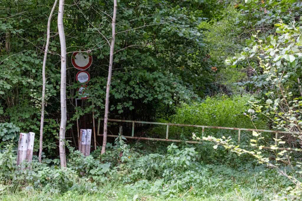 Ein verschlossenes Tor zugewachsen von diversen Pflanzen. Ein Straßenschild links am Bildrand