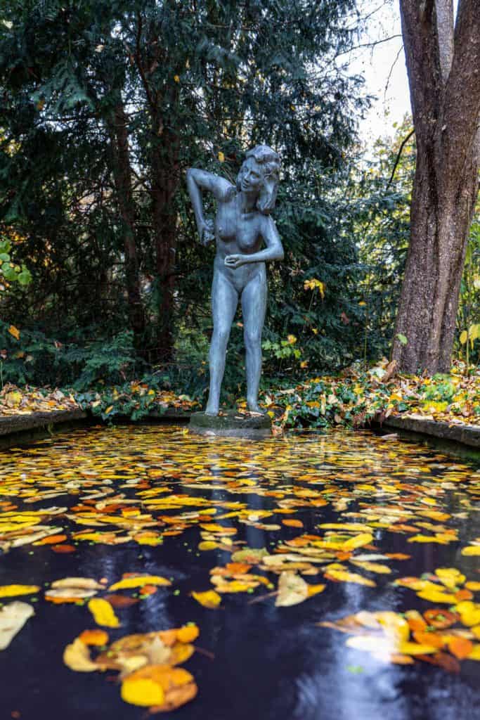 Unbekleidete weibliche Figur aus Bronze, die vor einem kleinen mit Wasser gefüllten Becken steht. Auf dem Wasser schwimmt Laub