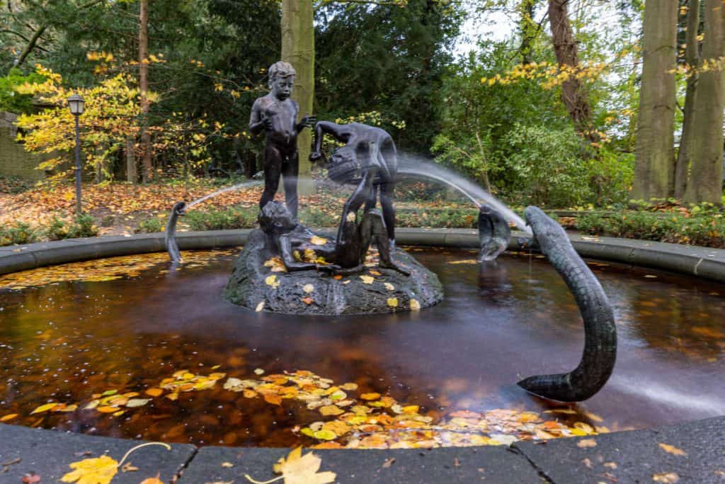 Brunnen in Langzeitbelichtung. In der Mitte drei Bronze Figuren spielender Kinder, außen drei Schlangen, die Wasser verspritzen