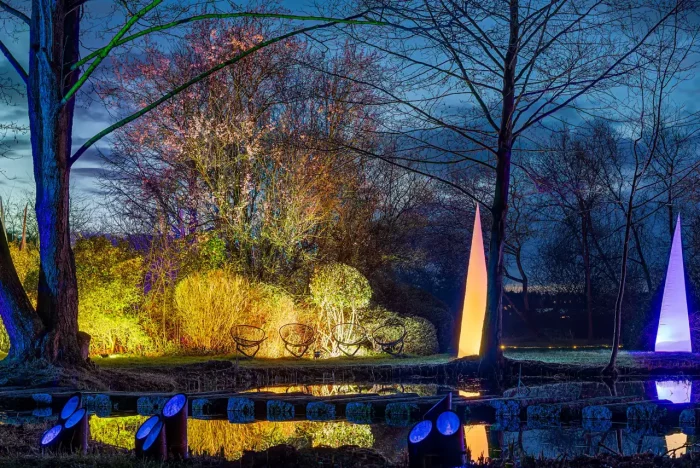 Nächtliche Ansicht eines Gartens mit farbenfroher Beleuchtung, darunter blau beleuchtete Objekte an einem Teich, konische Lichtskulpturen und in verschiedenen Farbtönen beleuchtete Bäume mit Spiegelungen im Wasser.