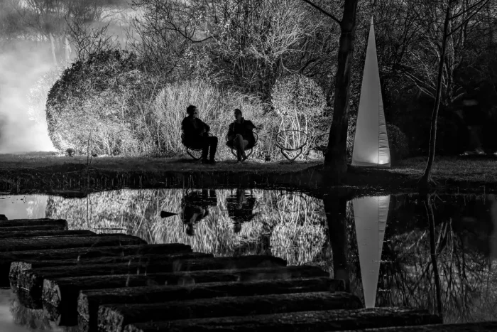 Ein Schwarz-Weiß-Bild, das zwei Personen zeigt, die auf verschiedenen Schaukeln in der Nähe eines Gewässers sitzen, mit einer reflektierenden Kunstskulptur und einer Reihe von Trittsteinen im Vordergrund.