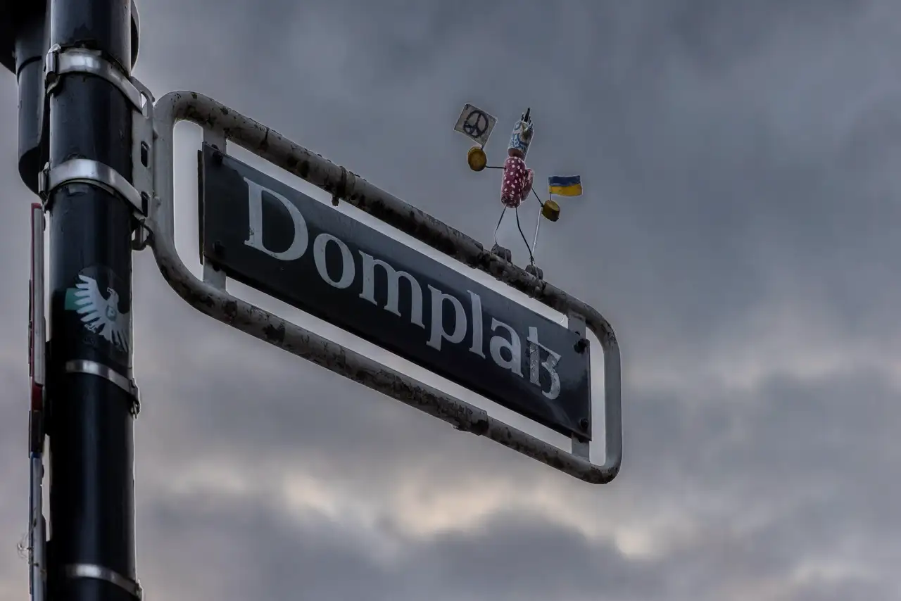Ein Straßenschild mit der Aufschrift "Domplatz" und der skurrilen Skulptur einer behelmten Figur, die ein Schild und Fahnen auf dem Schild hält, vor einem wolkenverhangenen Hintergrund.