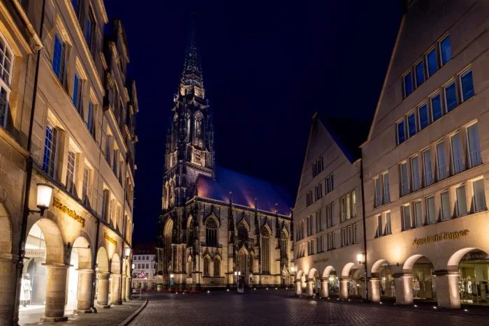 Ein nächtlicher Blick auf eine gotische Kathedrale mit einem hohen Turm neben modernen Gebäuden mit beleuchteten Schaufenstern auf einer Kopfsteinpflasterstraße.