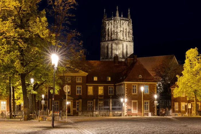 Eine nächtliche Ansicht eines historischen Platzes mit Kopfsteinpflaster, Straßenlaternen und einem großen, beleuchteten Kirchen- oder Kathedralenturm im Hintergrund, flankiert von Bäumen.