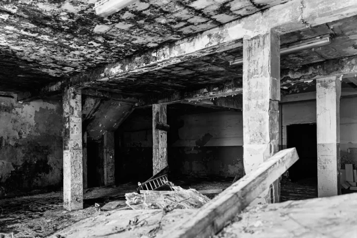 Schwarz-Weiß-Bild eines baufälligen Innenraums mit abblätternder Decke, bröckelnden Wänden und freiliegenden Betonbalken. Trümmer liegen auf dem Boden verstreut.