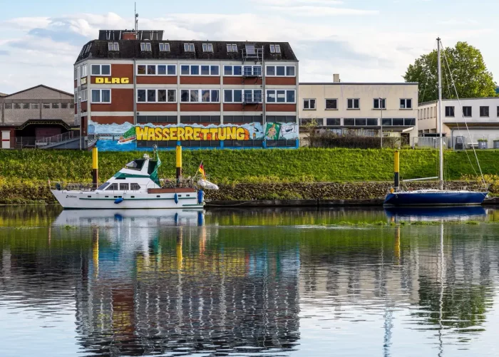 Eine Szene am Wasser mit zwei vertäuten Booten, einer weißen Motoryacht im Vordergrund und einem blauen Segelboot dahinter. Im Hintergrund ein mehrstöckiges Gebäude mit dem Schriftzug "DLRG WASSERRETTUNG" in großen Buchstaben und Grünflächen entlang der Uferlinie.
