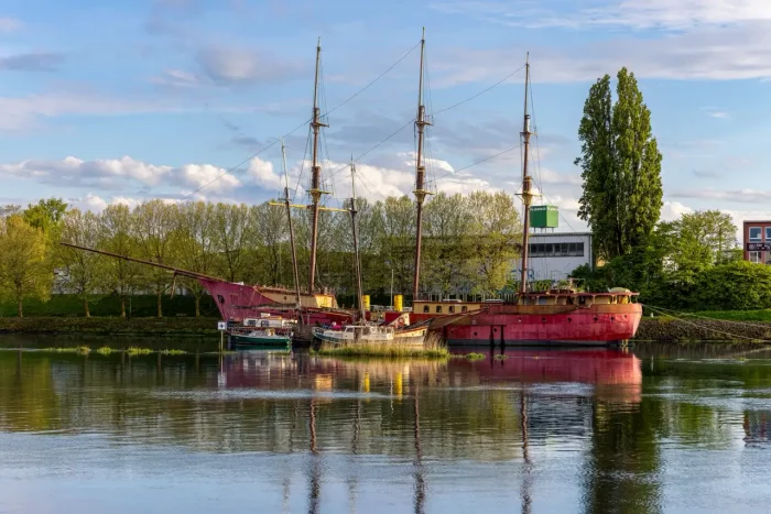Ein großes rotes dreimastiges Schiff, das an einem ruhigen Fluss festgemacht ist, mit Spiegelungen auf dem Wasser, grünen Bäumen und Gebäuden im Hintergrund unter einem blauen Himmel mit vereinzelten Wolken.