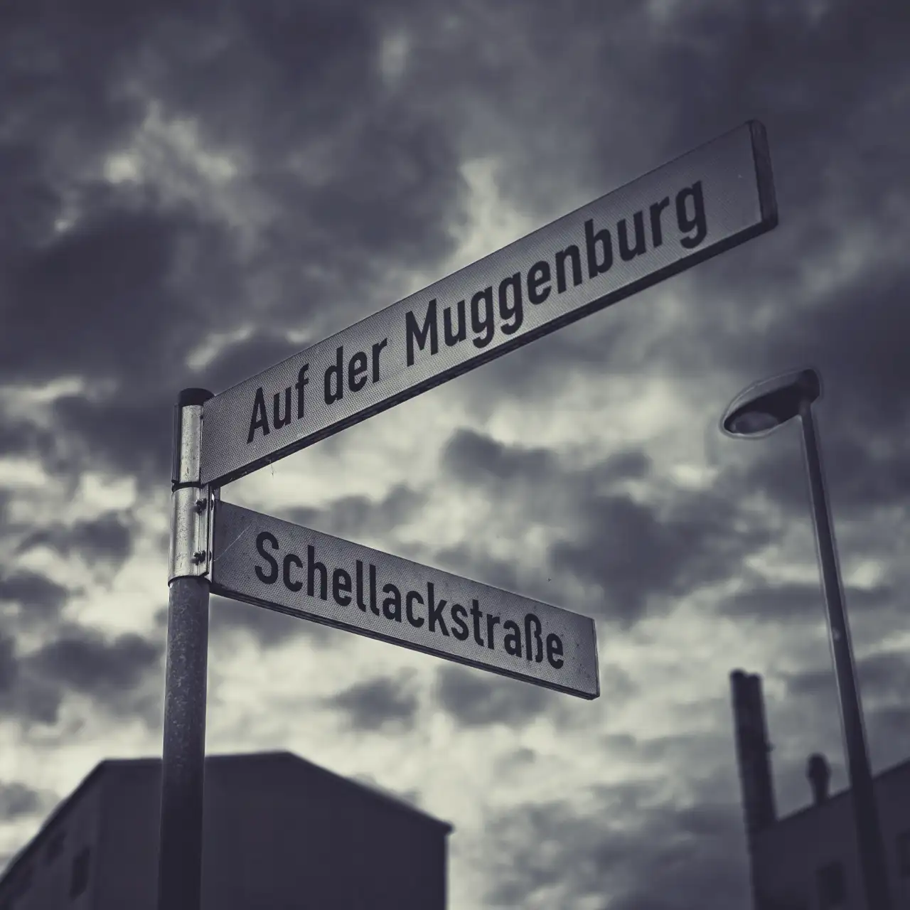 Straßenschilder "Auf der Muggenburg" und "Schellackstraße" an einer Kreuzung mit einem stürmischen Himmel im Hintergrund.