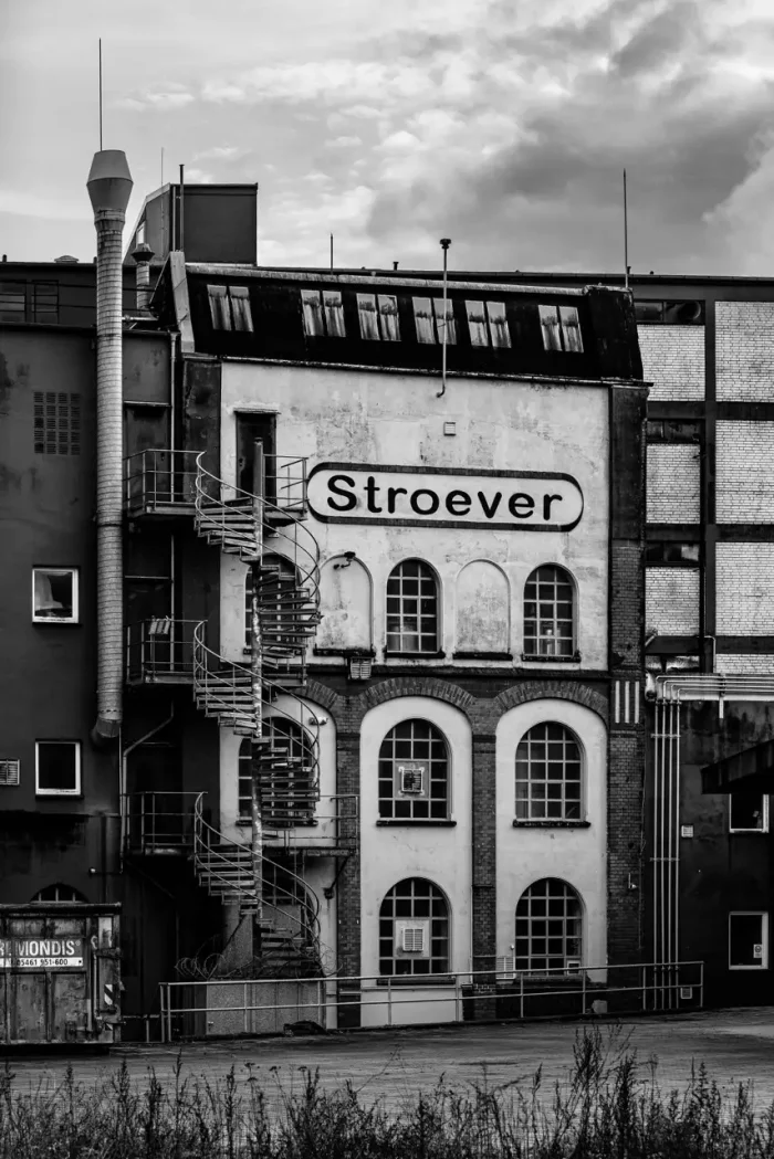 Schwarz-Weiß-Bild eines alten Industriegebäudes mit dem Namen "Stroever" an der Fassade, mit großen Bogenfenstern und einer äußeren Wendeltreppe.