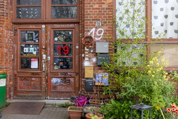 Eine verwitterte Tür eines Backsteingebäudes, bedeckt mit Graffiti, Aufklebern und Plakaten, mit einer großen roten Zahl "19" auf der Tür. Neben der Tür sind mehrere Briefkästen angebracht, und vor der Tür sind Topfpflanzen aufgestellt.