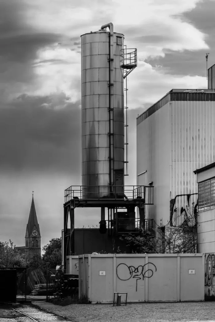 Schwarz-Weiß-Foto eines Industriesilos neben Gebäuden, mit einem Kirchturm im Hintergrund unter einem bewölkten Himmel.