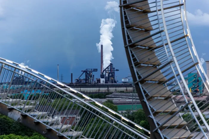 Industrieanlage, die Rauch emittiert, umrahmt von einem spiralförmigen Stahlweg im Vordergrund.