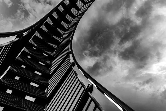 Schwarz-Weiß-Foto einer geschwungenen Treppe von unten vor einem bewölkten Himmelshintergrund.