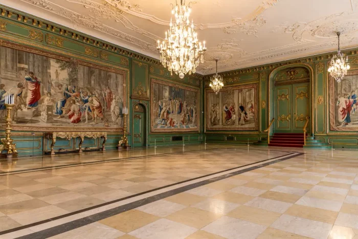 Eine verzierte, geräumige Halle mit Kronleuchtern, detaillierten Deckenleisten, großen Wandteppichen, die historische Szenen darstellen, einer grünen und goldenen Wanddekoration und einem polierten karierten Marmorboden.