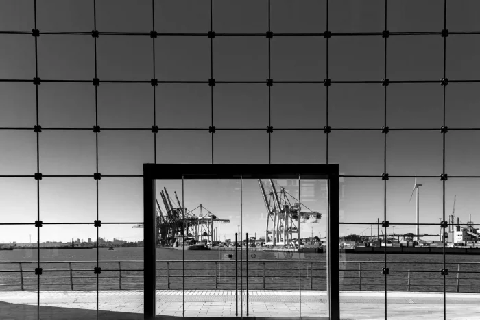 Monochromes Bild eines Hafens mit Kränen, gesehen durch ein großes Glasfenster, eingerahmt von einem Gitter aus dunklen Pfosten.