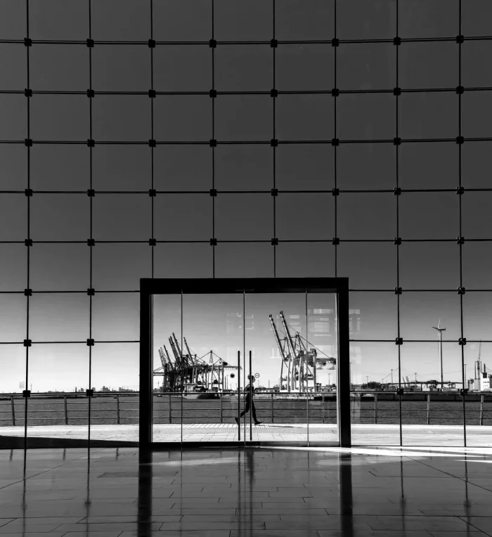 Monochromes Bild eines Hafens mit Kränen, gesehen durch ein großes Glasfenster, eingerahmt von einem Gitter aus dunklen Pfosten.