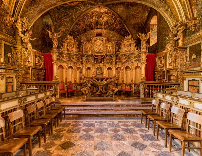 Geschmückte, historische Kapelle mit komplizierten goldenen Dekorationen, an den Seiten gesäumten Holzstühlen, roten Vorhängen und detaillierten Wandmalereien an den Wänden und der Decke.