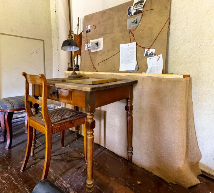 Ein hölzerner Schreibtisch und Stuhl in einem Raum mit einer Pinnwand an der Wand, die mit Papieren, Fotos und einer roten Schnur bedeckt ist, die verschiedene Punkte verbindet. Eine Lampe sitzt auf dem Schreibtisch.