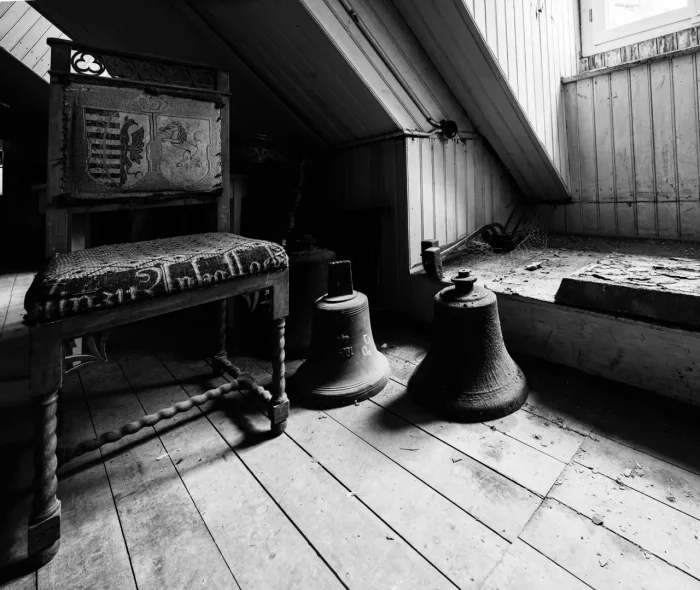 Schwarz-Weiß-Foto eines Dachbodens mit einem Holzstuhl mit einer Tapisserie-Rückenlehne, zwei großen Glocken auf einem Holzboden und einem Fenster, das Licht hereinlässt.