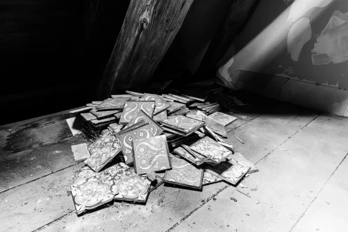 Ein Haufen dekorativer Fliesen mit komplizierten Mustern, verstreut auf einem verlassenen Boden mit Holzbrettern, teilweise vom Sonnenlicht beleuchtet.