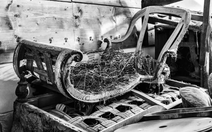 Schwarz-Weiß-Bild eines teilweise zerlegten und beschädigten antiken Sofas mit freiliegenden Federn und Holzrahmen. Der Hintergrund zeigt Holzbretter und andere alte Möbelstücke.