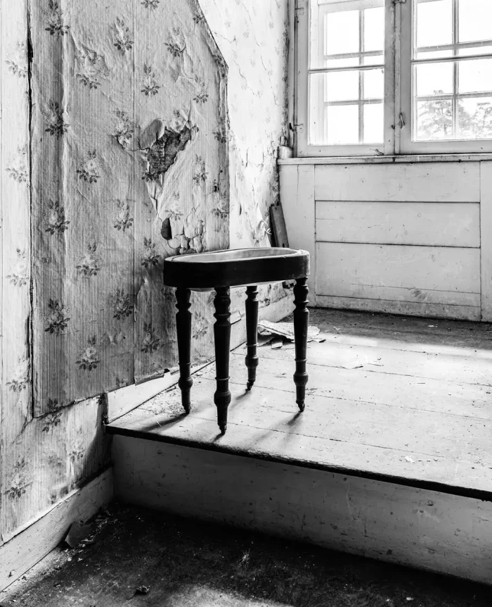 Schwarz-Weiß-Fotografie eines alten, maroden Innenraums mit abblätternden Tapeten und einem kleinen Holzhocker. Sonnenlicht scheint durch ein Fenster und wirft Schatten auf den Boden.