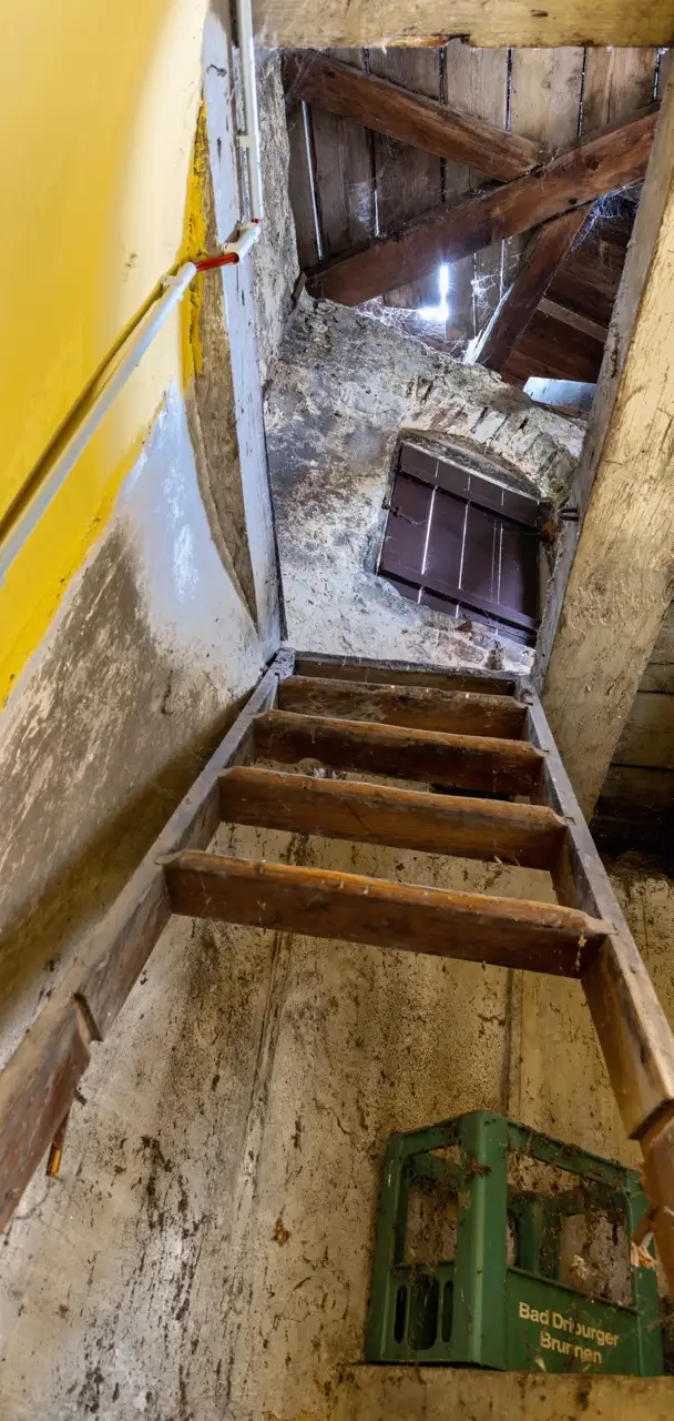 Ein Blick auf eine verfallene Holztreppe, die zu einem Dachboden mit freiliegenden Balken führt. Die Wände und die Decke sind verwittert, und unten befindet sich eine grüne Kiste mit der Aufschrift "Bad Driburger Brunnen".