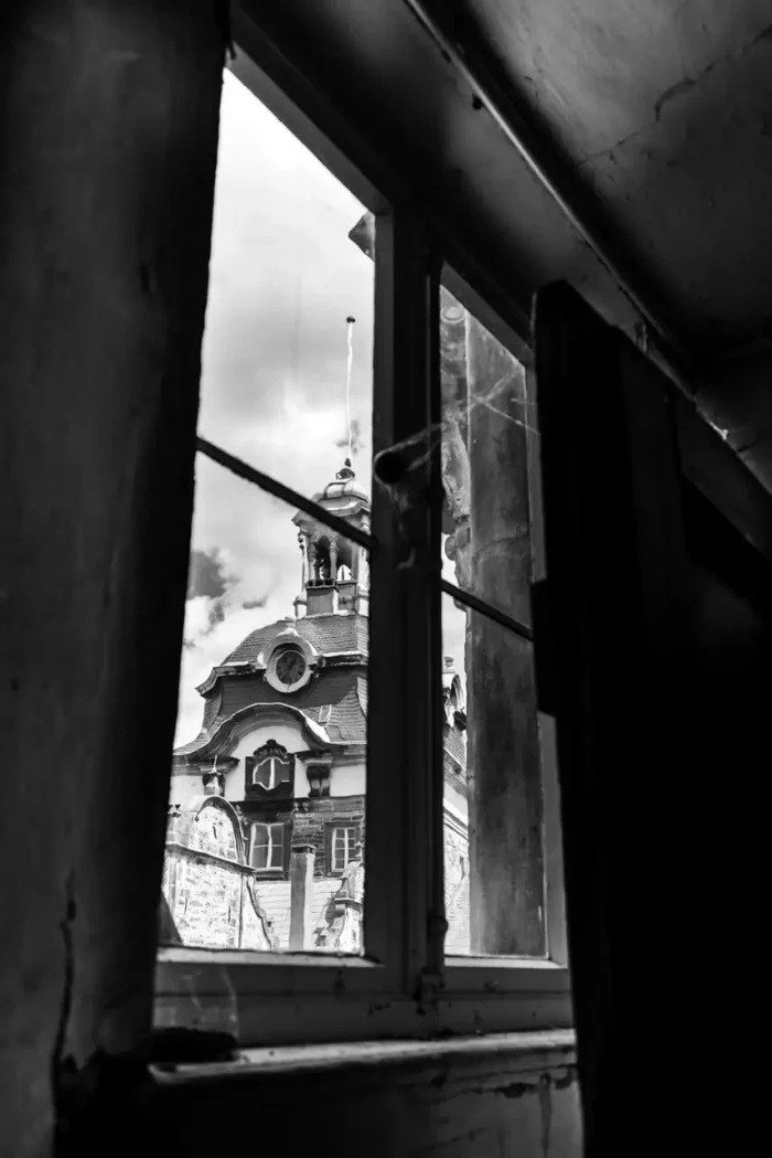 Blick auf ein altes Gebäude mit einer verzierten Fassade, durch ein Fenster aus dem Inneren eines dunklen Raumes gesehen.