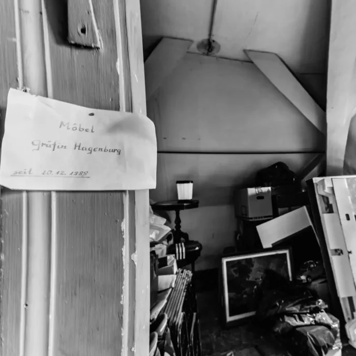 Schwarz-Weiß-Bild eines überladenen Dachbodens mit einem Schild an der Tür mit der Aufschrift "Möbel Gräfin Hagenburg seit 20.12.1988". Der Dachboden enthält verschiedene Kisten, ein Gemälde und alte Möbel.