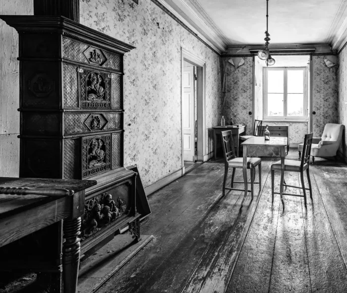 Schwarz-Weiß-Fotografie eines alten, spärlich eingerichteten Raumes mit Holzböden. Auf der linken Seite befindet sich ein dekorativer Holzofen, ein Tisch mit drei Stühlen in der Mitte, ein Beistelltisch und ein Stuhl am Fenster und gemusterte Tapeten.