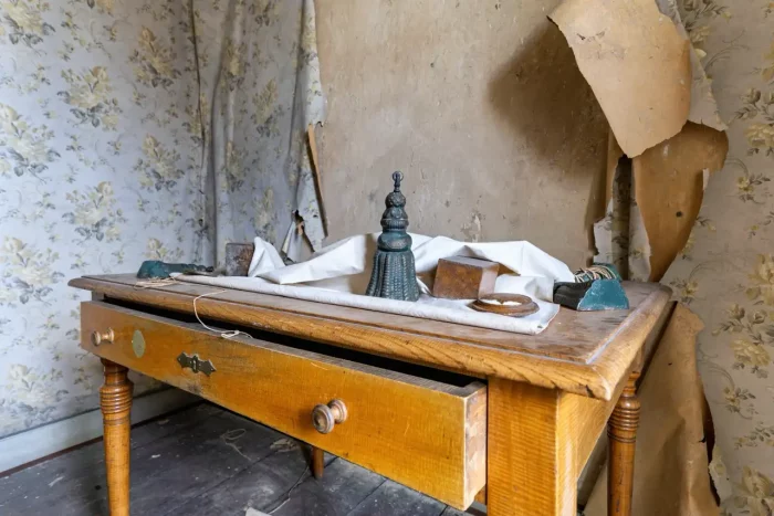 Ein alter Holztisch mit einer offenen Schublade, der verschiedene Objekte wie einen kleinen dekorativen Gegenstand und ein Tuch vor einem Hintergrund aus zerrissenen Blumentapeten zeigt.