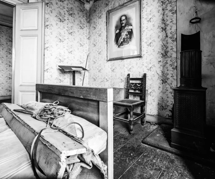 Ein Schwarz-Weiß-Bild eines Vintage-Zimmers mit einem Holzbett, einem Stuhl, einem gerahmten Porträt an der Wand, einem kleinen Rednerpult und alten Tapeten. Der Boden erscheint abgenutzt, und es gibt Unordnung auf dem Bett.