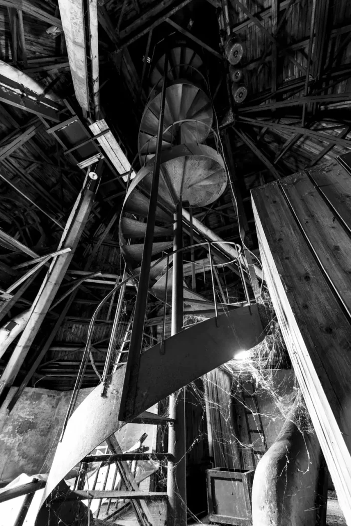 Schwarz-Weiß-Bild einer alten Wendeltreppe in einem verfallenen Gebäude mit Spinnweben und einem verwitterten, hölzernen Interieur.