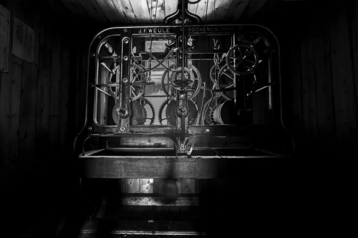 Ein Schwarz-Weiß-Bild eines alten mechanischen Uhrenmechanismus mit Zahnrädern und Zahnrädern vor einem Holzhintergrund. Die Maschine ist mit "J.F. Weule Bockenem 1896" beschriftet. Der Raum ist dill beleuchtet und wirft Schatten um das Werk.