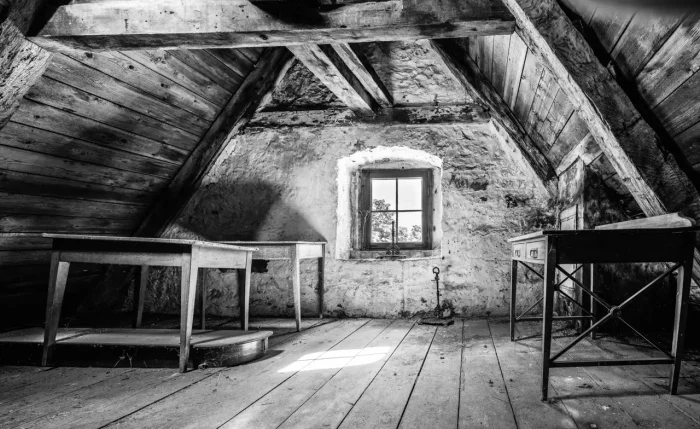 Ein Schwarz-Weiß-Foto eines alten Dachbodenzimmers mit Holzbalken, zwei kleinen Tischen, einem Fenster und einer freiliegenden Backsteinmauer.