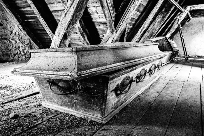 Schwarz-Weiß-Foto eines Dachbodens mit drei alten, verzierten Holzsärgen, die mit Staub und Spinnweben bedeckt sind.