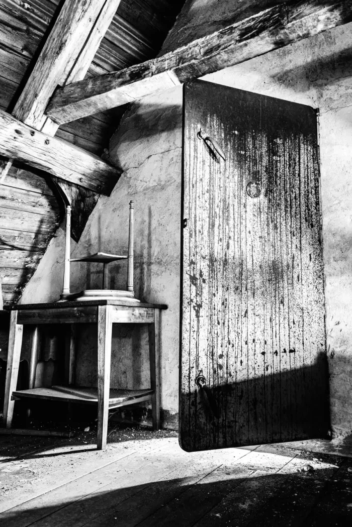 Schwarz-Weiß-Bild eines alten, rustikalen Dachbodens mit einer verwitterten Tür leicht offen und einem kleinen Tisch mit umgedrehten Stühlen darauf. Die Wände und Holzbalken sind abgenutzt und gealtert.