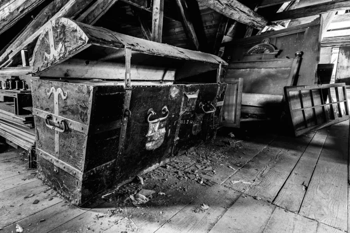Eine alte, offene Truhe auf einem Dachboden mit verstreuten Trümmern und Möbelstücken in der Nähe. Schwarz-Weiß-Foto.