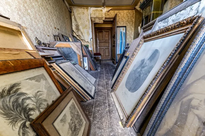 Ein überladener Raum mit mehreren geneigten gerahmten Gemälden und Kunstwerken, verzierten Tapeten und einer Holztür im Hintergrund. Die Wände zeigen Anzeichen von Abblättern und Beschädigung.
