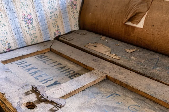 Alte Holzkiste mit der Beschriftung "MILITAIRE BAKKERY", mit einer verwitterten Verriegelung, die auf einem Vintage-Blumentapetenhintergrund gesetzt ist.