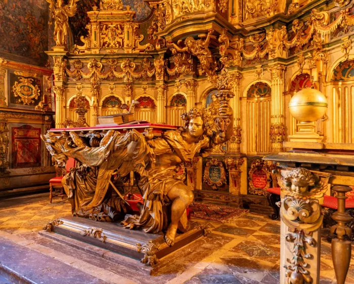 Aufwendiger goldener Altar mit detaillierten Schnitzereien und einer zentralen Statue eines knienden Engels, der eine Plattform mit einem Buch stützt.