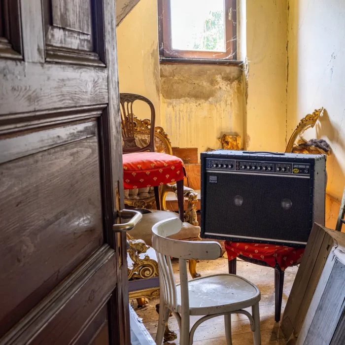 Ein überladener Raum mit einer offenen Holztür, mehreren verzierten Stühlen mit roter Polsterung, einem weißen Stuhl und einem Gitarrenverstärker. Die Wände schälen sich und der Raum scheint vernachlässigt zu sein.