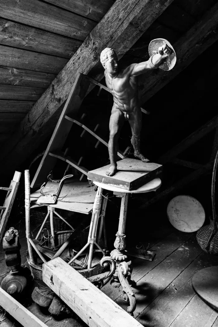 Ein Schwarz-Weiß-Bild eines überladenen Dachbodens mit einer alten Statue eines nackten Mannes, der ein Schild hält, auf einem kleinen Tisch inmitten verstreuter Möbel und Gegenstände.