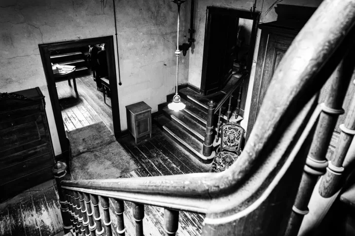 Schwarz-Weiß-Bild einer alten, schmalen Treppe, die zu verschiedenen Türen in einem dunkel beleuchteten, rustikalen Interieur mit Holzböden und antiken Möbeln führt.