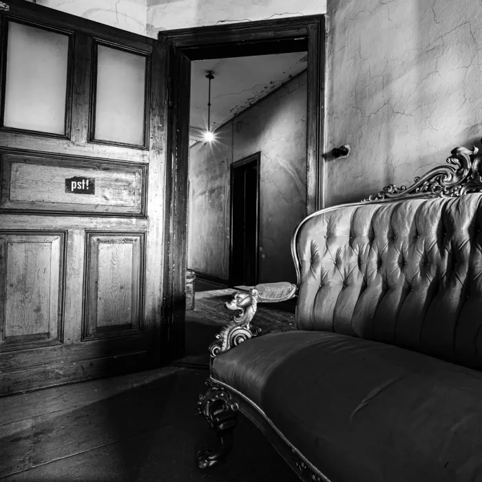 Ein Schwarz-Weiß-Foto eines Innenraums mit einem verzierten, getufteten Sofa und einer teilweise offenen Tür auf der "pst!" steht. Über der Tür ist ein schumrig beleuchteter Flur sichtbar.