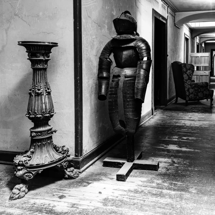 Schwarz-Weiß-Foto eines Innenflurs mit einer Rüstung auf einem Ständer, einem verzierten Sockel und einem Stuhl mit Blumenmuster im Hintergrund. Der Flur erscheint gealtert mit sichtbarer Abnutzung auf dem Holzboden.