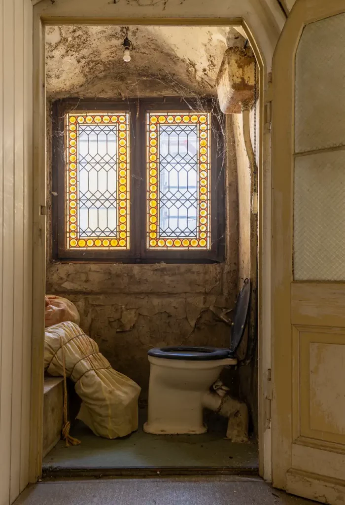 Ein altes, düster beleuchtetes Badezimmer mit Buntglasfenstern, abblätternden Wänden, einer Toilette mit einem schwarzen Sitz, einer Glühbirne, die von der Decke hängt, und etwas Unordnung auf dem Boden.